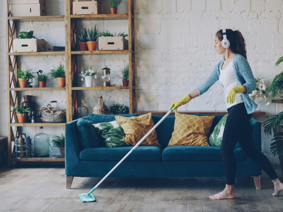 Floor Care Schedule For Cleaner, Longer-Lasting Floors 1 Woman wearing headphones and rubber gloves uses a mop to clean the floor in a living room with a teal sofa, plants, and shelves in the background, sticking to her regular floor care schedule.