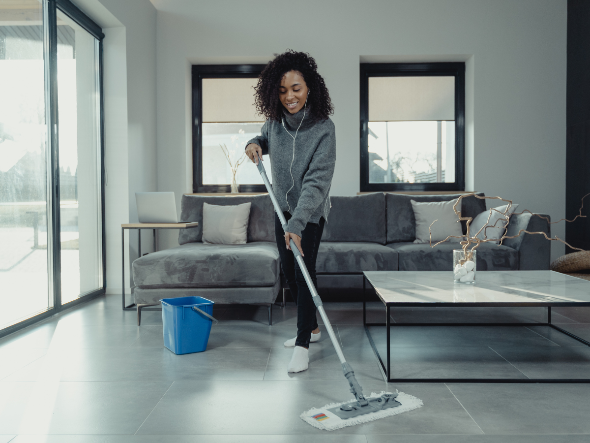 Floor Care Schedule For Cleaner, Longer-Lasting Floors 3 Woman wearing headphones mops the floor in a modern living room, following her regular floor care schedule, with a blue bucket nearby and sunlight streaming through large windows.