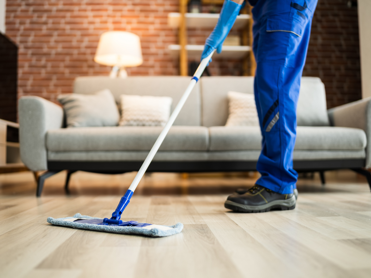Person in blue work clothes and gloves mopping a wooden floor as part of a regular floor care schedule in a living room with a gray sofa, pillows, lamp, and brick wall in the background.