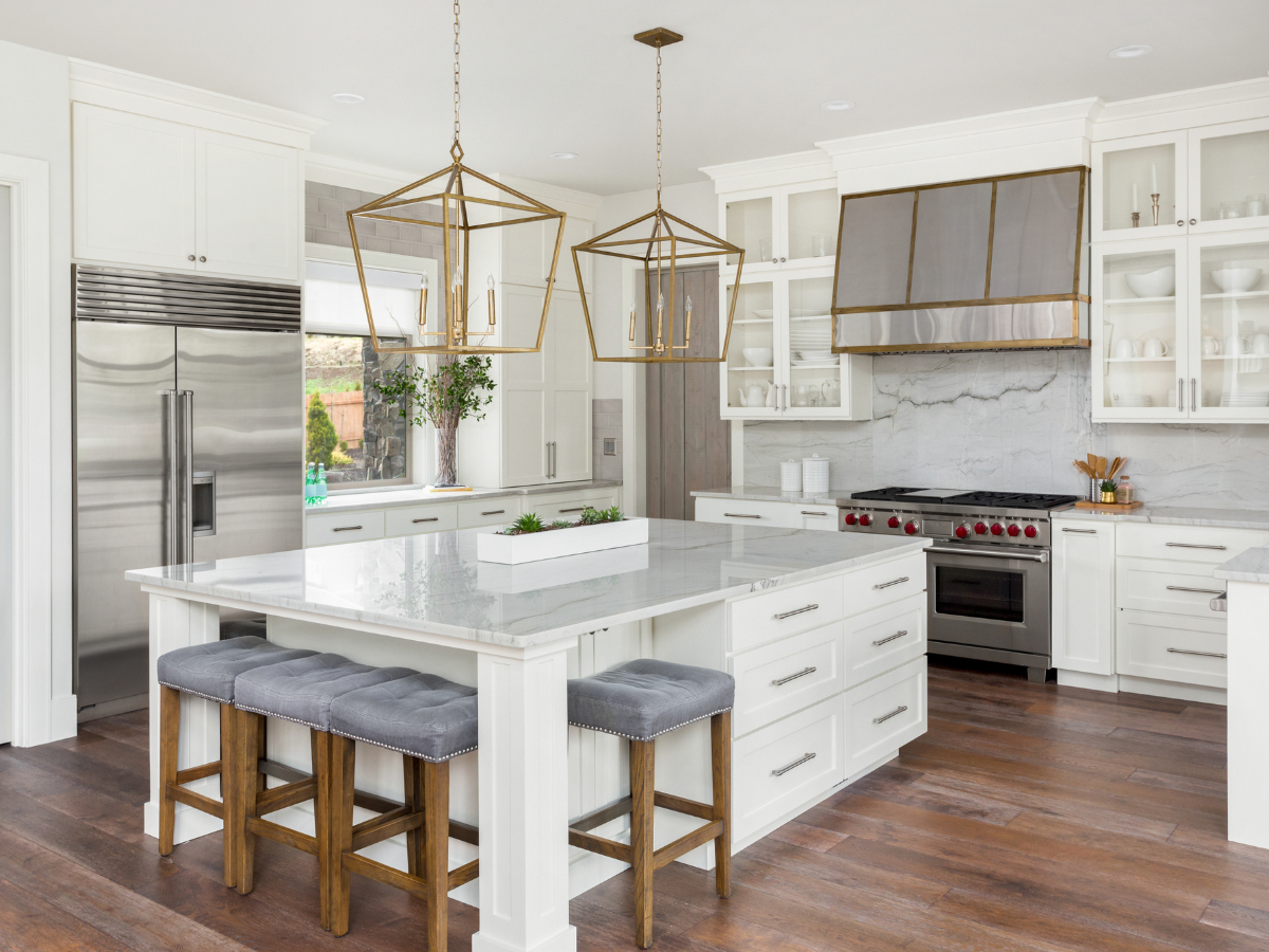 Modern kitchen with white cabinets, marble countertops, stainless steel appliances, two pendant lights, a large island with gray cushioned stools, and elegant wood flooring&mdash;perfect for those considering tile vs hardwood for their own kitchen remodel.
