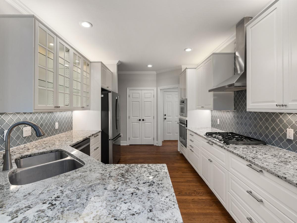 Modern kitchen with white cabinets, stainless steel appliances, granite countertops, a double sink, and a blue tile backsplash. Highlighting the appeal of tile vs hardwood surfaces, this space features beautiful hardwood floors complemented by recessed lighting.