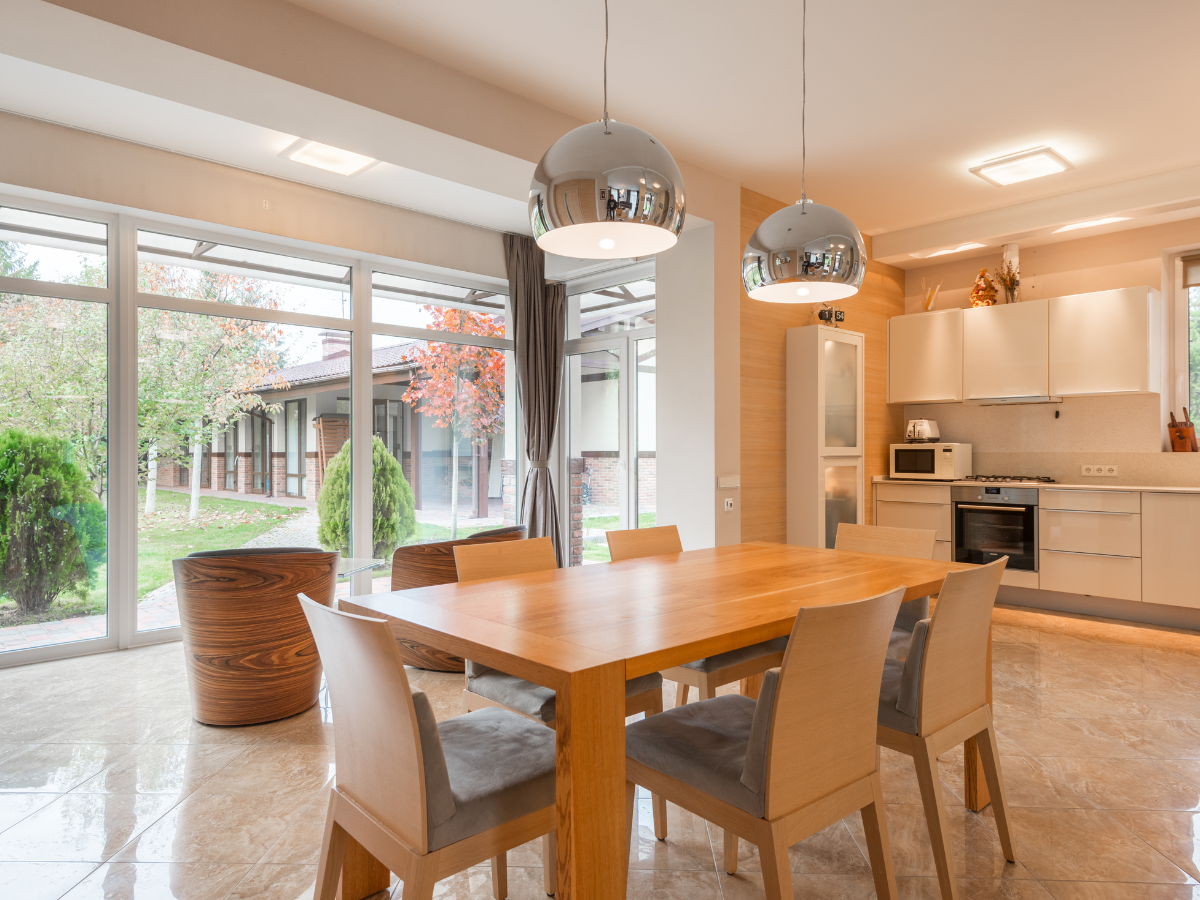 Modern kitchen and dining area with a wooden table, six chairs, pendant lights, and large windows overlooking a garden&mdash;perfect for those considering the tile vs hardwood debate when choosing stylish and practical flooring.
