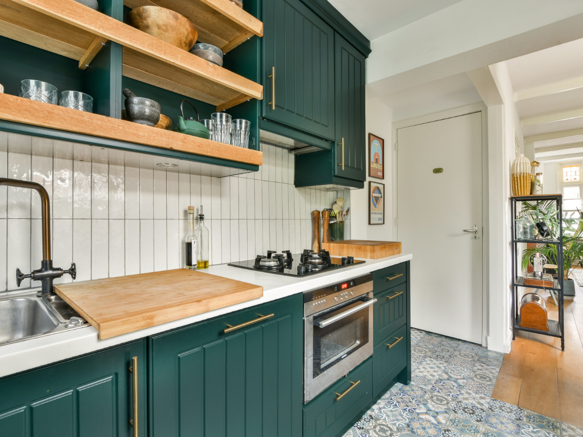 Modern kitchen with dark green cabinets, open wooden shelves, tiled backsplash, built-in oven, gas stove, and patterned floor tiles. Natural light brightens the space and highlights the elegant patterned tiles&mdash;an excellent choice for those considering tile vs hardwood for their kitchen floors.