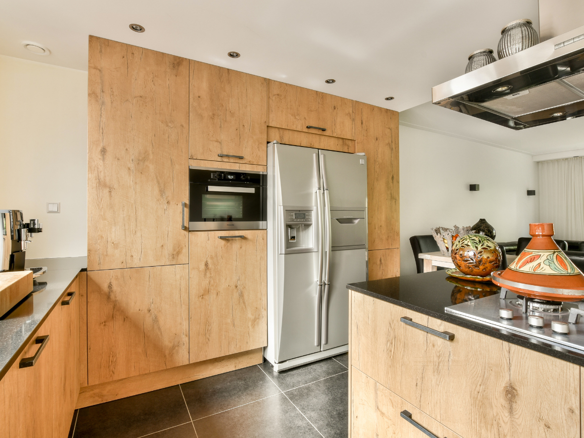 Modern kitchen with light wood cabinets, a built-in oven, stainless steel double-door fridge, black countertops, and a gas stove with cookware on the island&mdash;perfect for anyone debating tile vs hardwood flooring in their home.