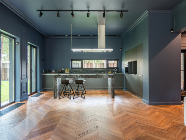 Modern kitchen with blue walls, marble island, two stools, and wooden herringbone floor. Large windows provide natural light and a view of the backyard—a stylish space that highlights the timeless appeal of hardwood when comparing tile vs hardwood flooring options.