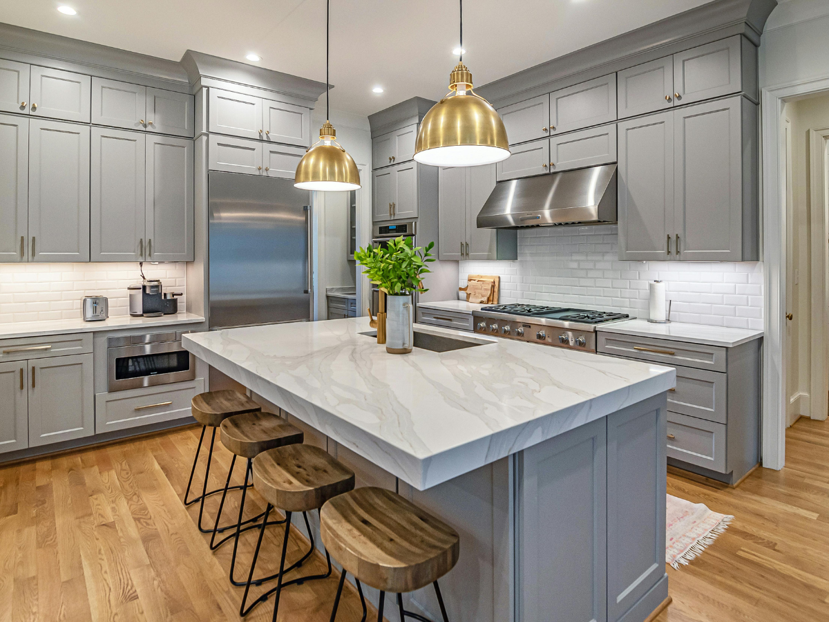Modern kitchen with gray cabinets, stainless steel appliances, white marble island with wooden stools, pendant lights, and gorgeous hardwood floors&mdash;an inviting space that highlights the timeless appeal of hardwood in the tile vs hardwood debate.